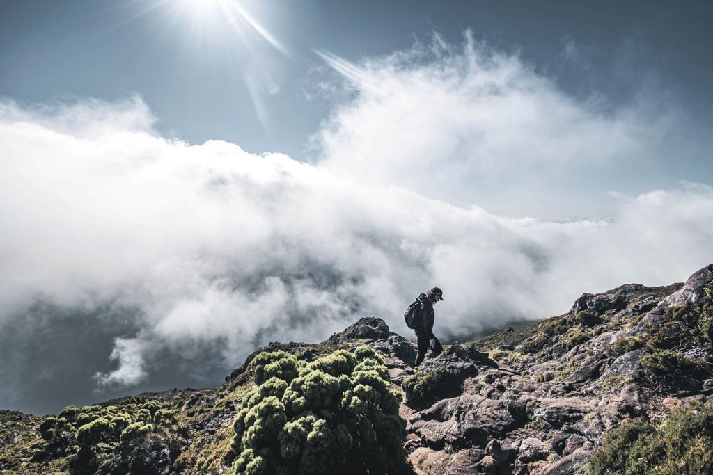 A lone hiker ascends a rocky mountain amidst clouds under a bright sun.