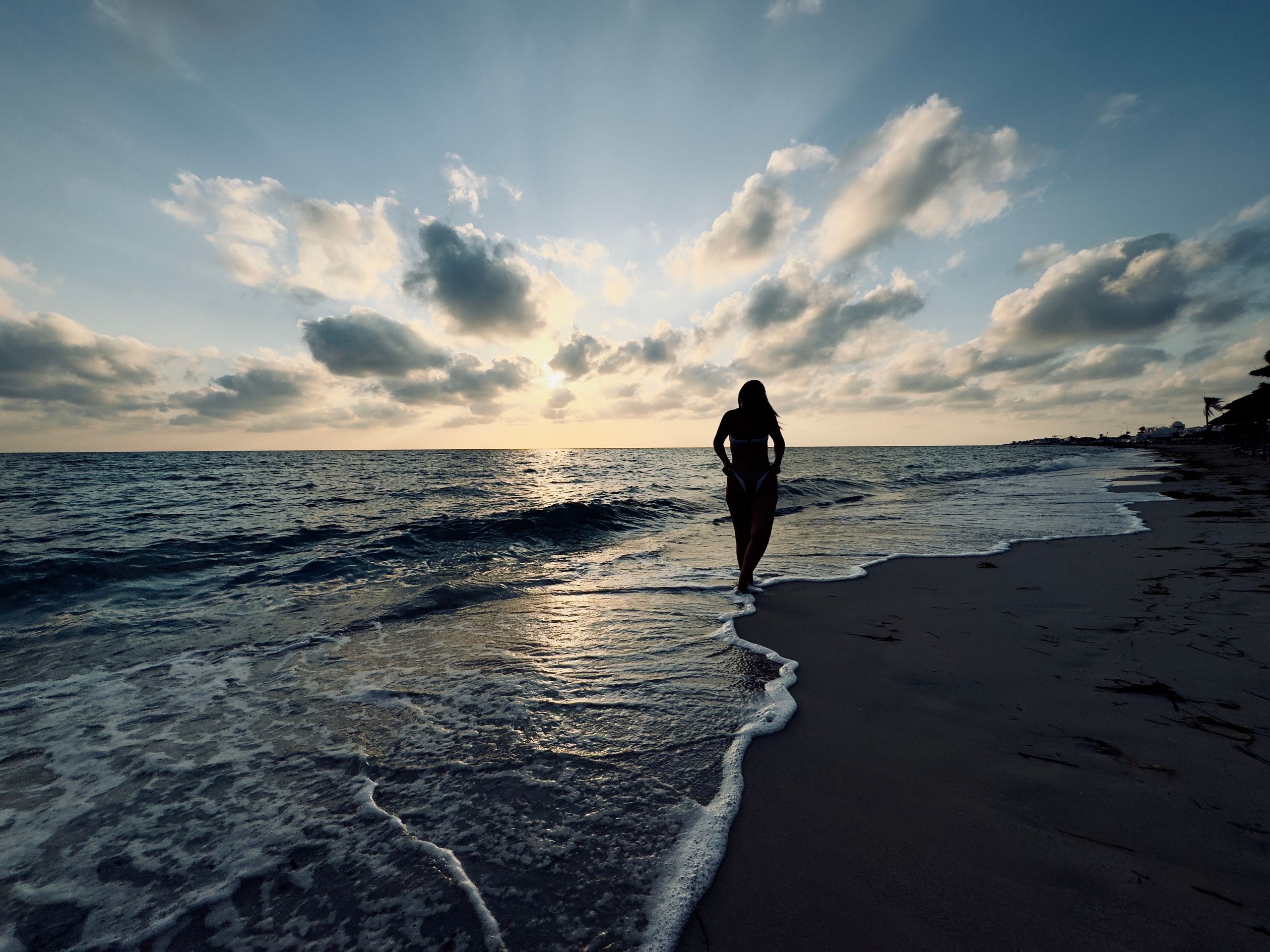 Captivating silhouette of a woman strolling along a Tunisian beach at sunset, with dramatic clouds and waves.